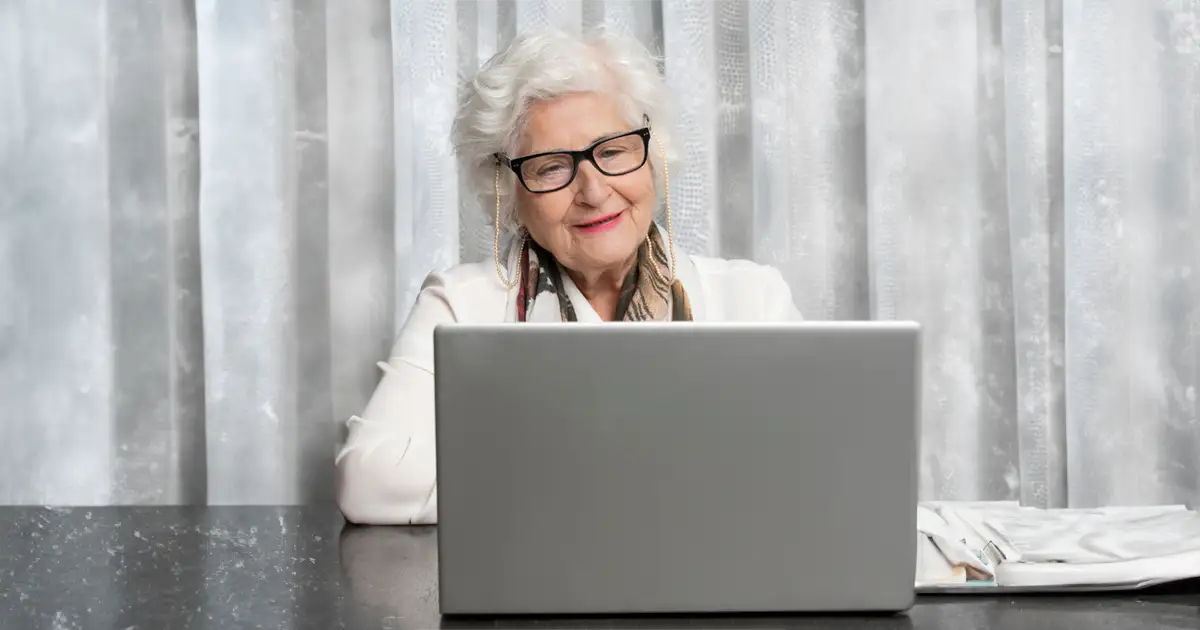 An elderly lady is sitting in front of a laptop, smiling