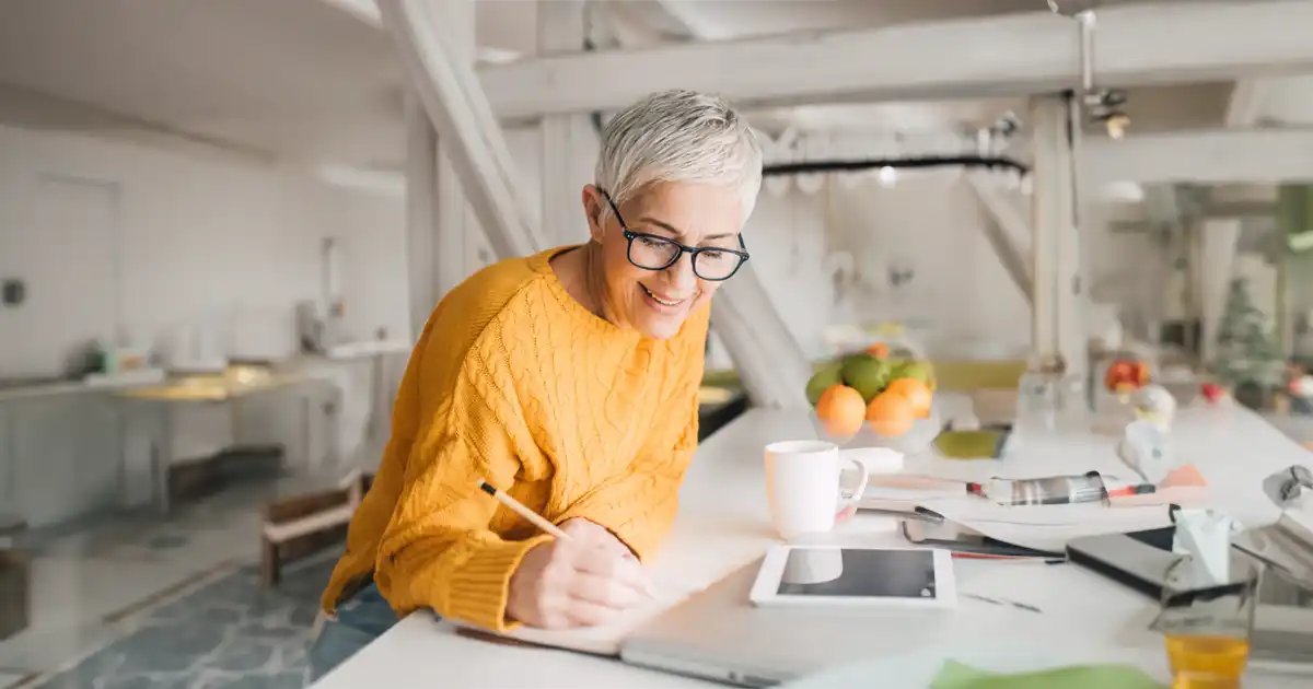 A mature woman is sitting at her desk with a laptop and iPad while she writes in her journal