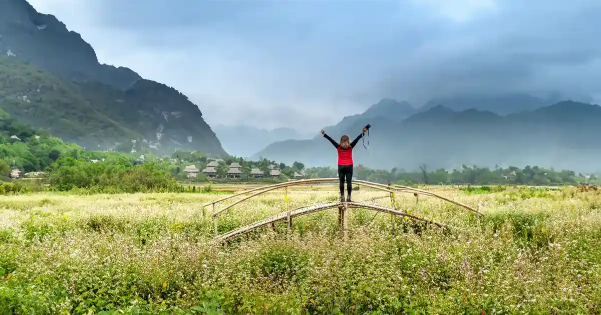 A woman standing on a bridge with her arms in the air in celebration, looking at spectacular mountains in the distance