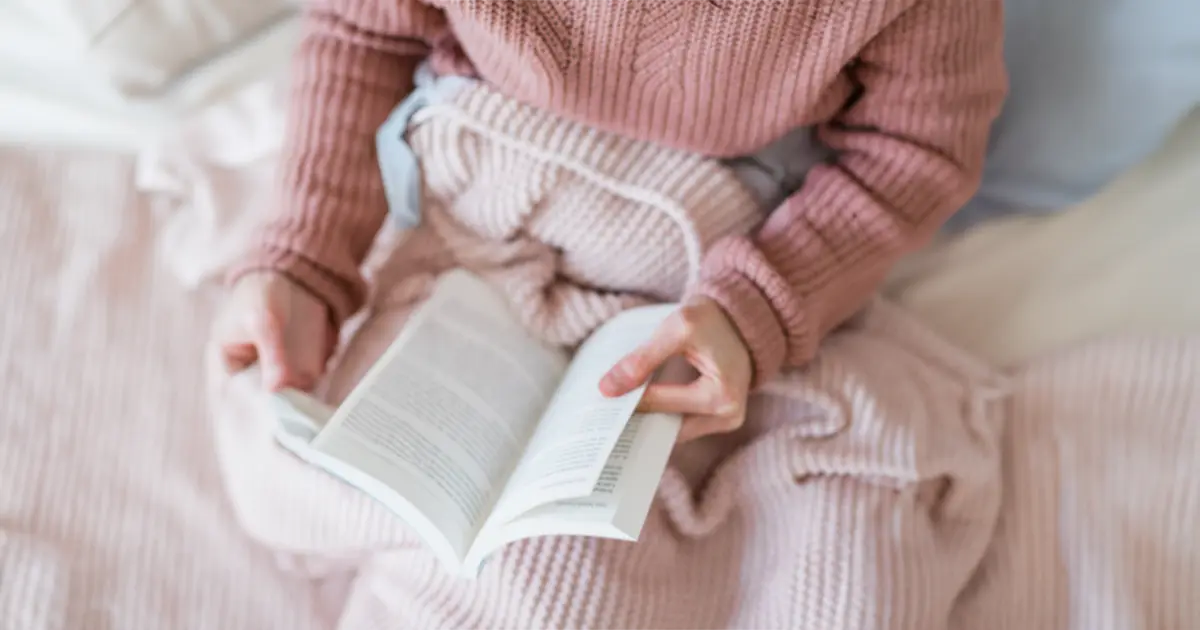 A woman from the torso down, is sitting cross legged, snuggled in pink blankets reading a book