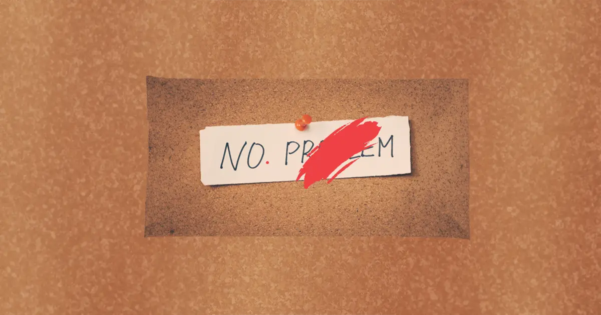 The words no problem are pinned on a cork board. The word problem has been crossed out in red paint, so the note now says no, with a red full stop after it.