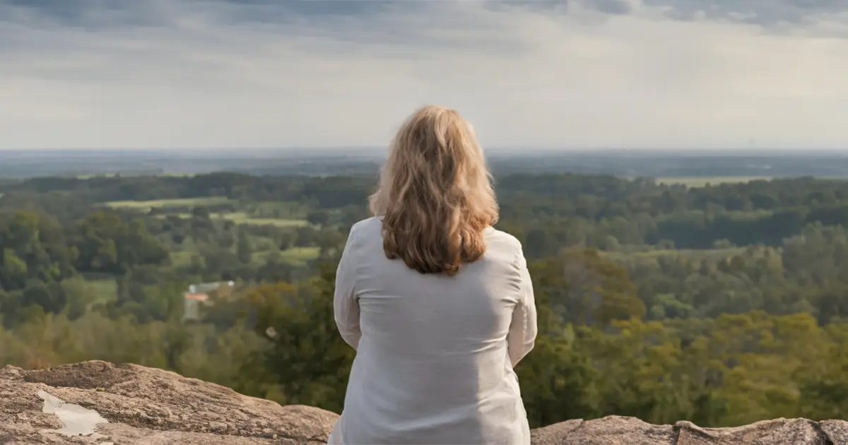 A woman is sitting on rocks with her back to us, as she looks at the valley vista below her