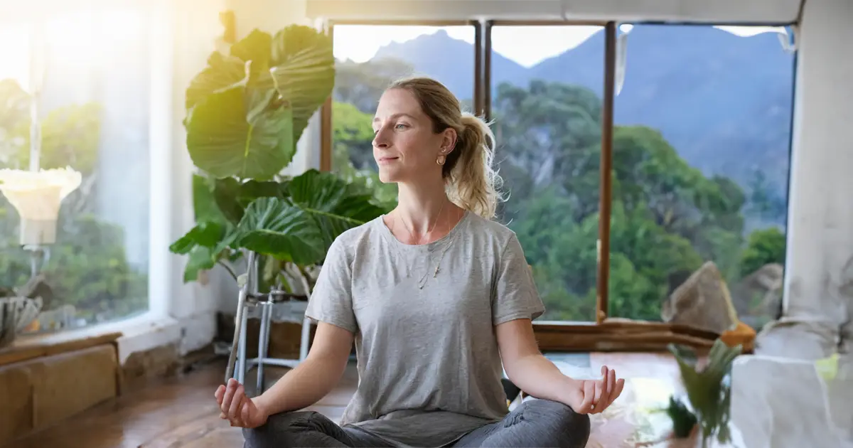 A woman with light hair is meditating while looking serenely out the window