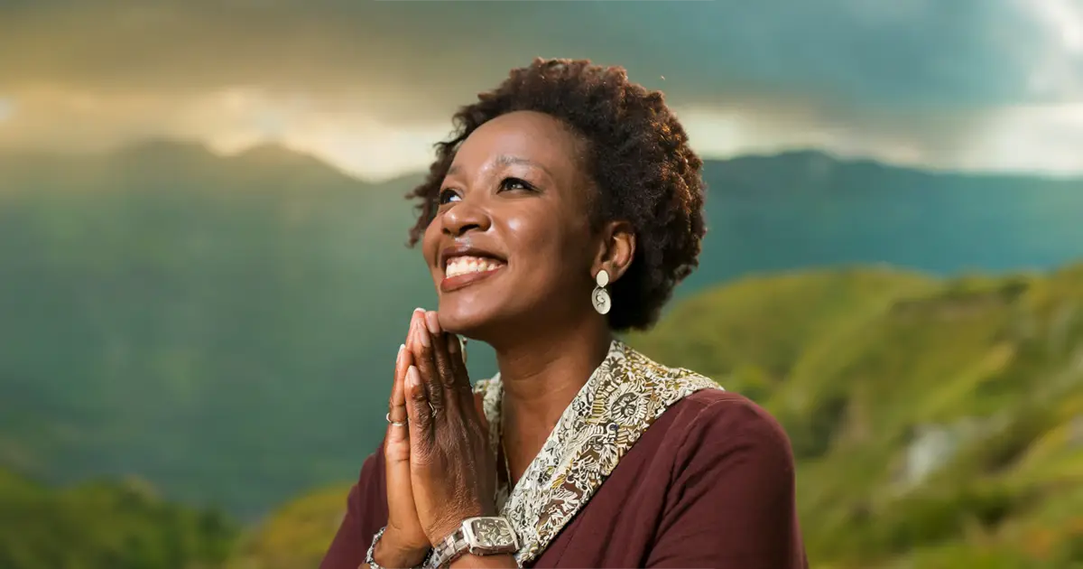 A woman with dark curly hair is praying while looking happily at the sky, she is also surrounded by lush mountains