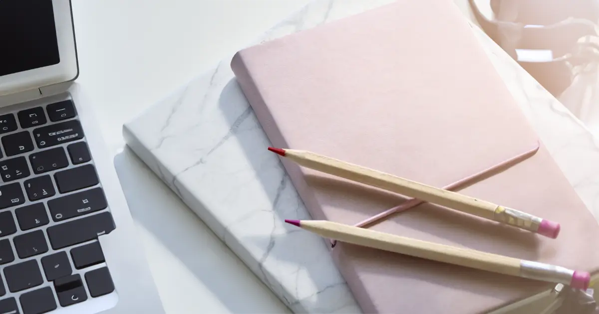 A pink journal sits atop another journal in marble style covering. A red pencil and purple pencil sit atop the journals.