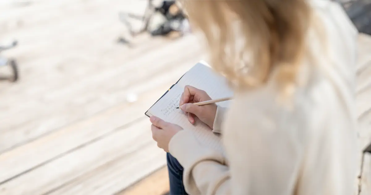 A woman is sitting down and writing in her journal with a coloured pencil