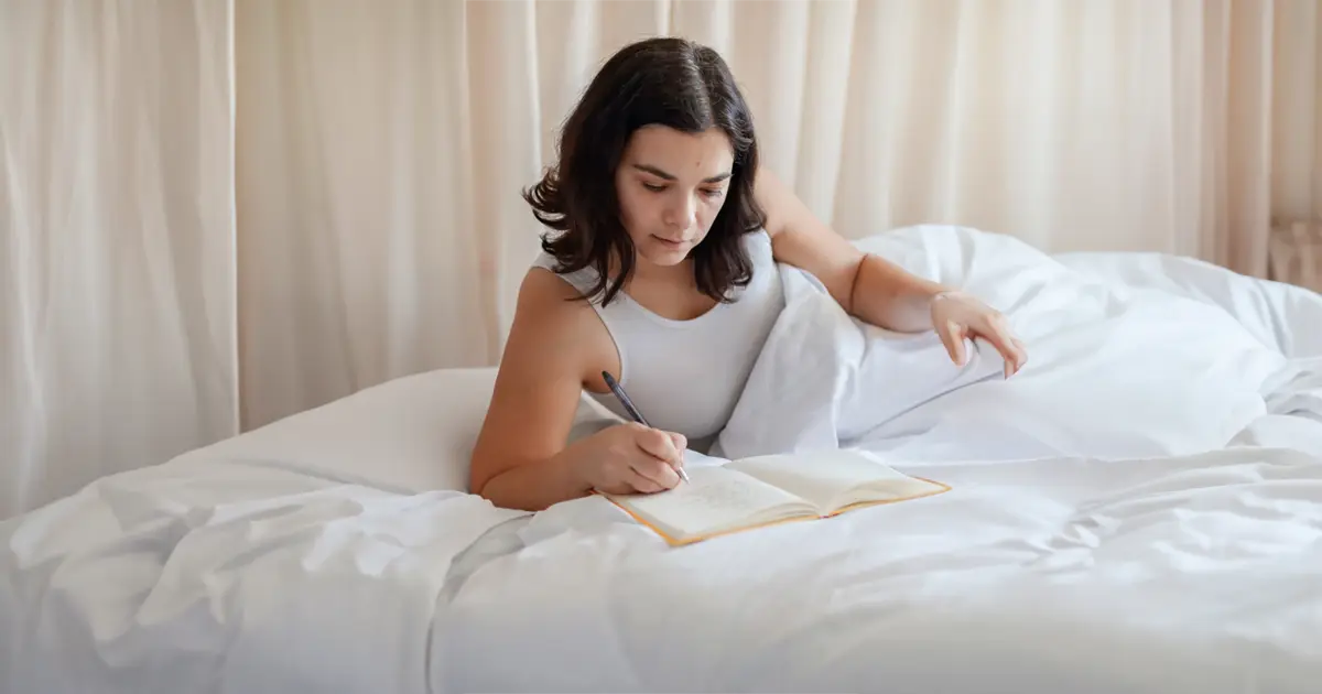 A woman is lying on a bed with a white curtain behind her while she writes in her journal