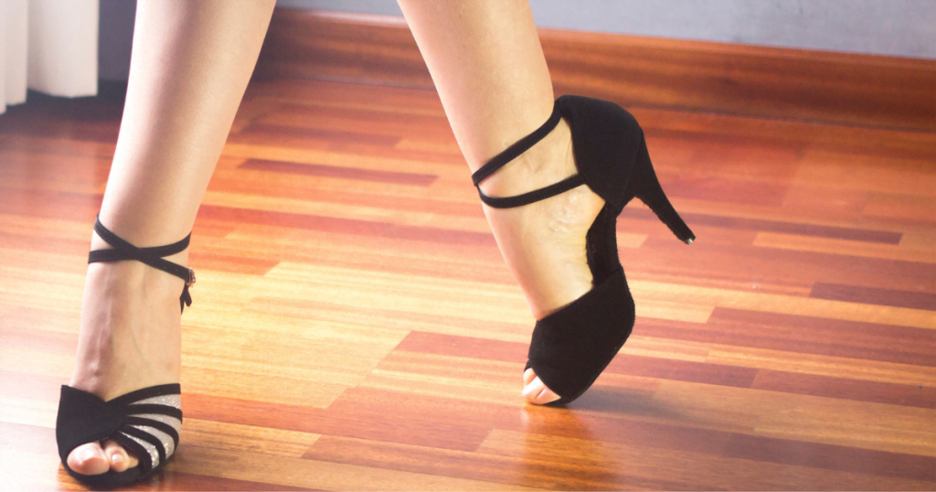 A woman photographed from the ankles down, striking a pose in black strappy ballroom dancing shoes. 