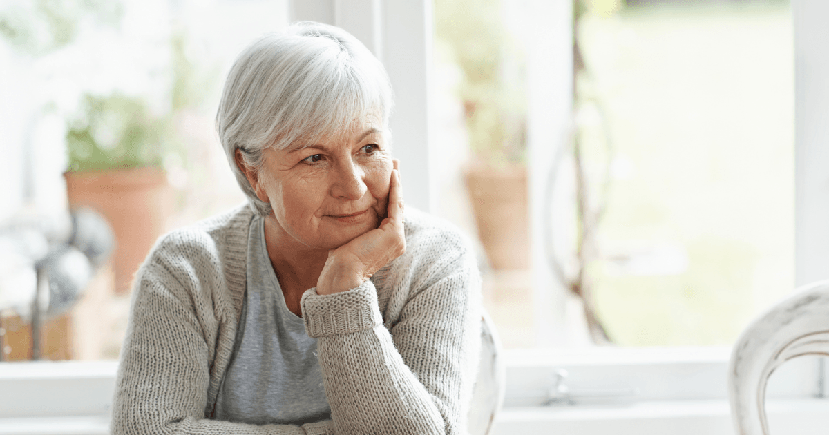 A grey haired woman sits at a table looking thoughtful, with her head resting on her chin.